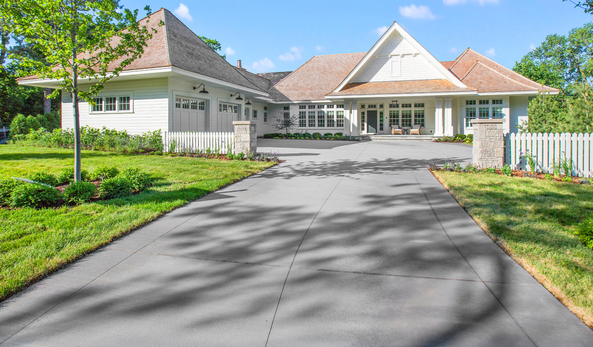 Home with a large driveway, white fence, and pitched roofs surrounded by a landscaped lawn and trees under a blue sky.