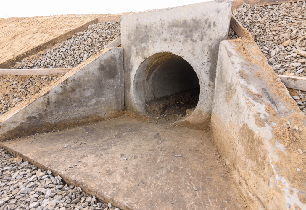 A concrete drainage culvert, reminiscent of a hidden home for small creatures, is surrounded by gravel and earth, featuring an open circular entrance.